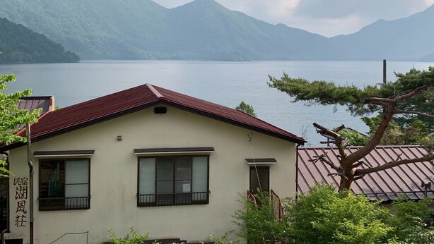 A building with a red roof and a sign in a foreign language, overlooking a calm lake with mountains in the background.