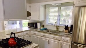 Una cocina con armarios blancos, un refrigerador de acero inoxidable, un microondas, un lavamanos y una tetera roja.