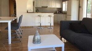 Ein modernes Wohnzimmer mit einer Couch, einem Esstisch mit Stühlen, einer Küchenzeile mit weißen Schränken und einer silbernen Dekorationsvase auf einem Tisch.