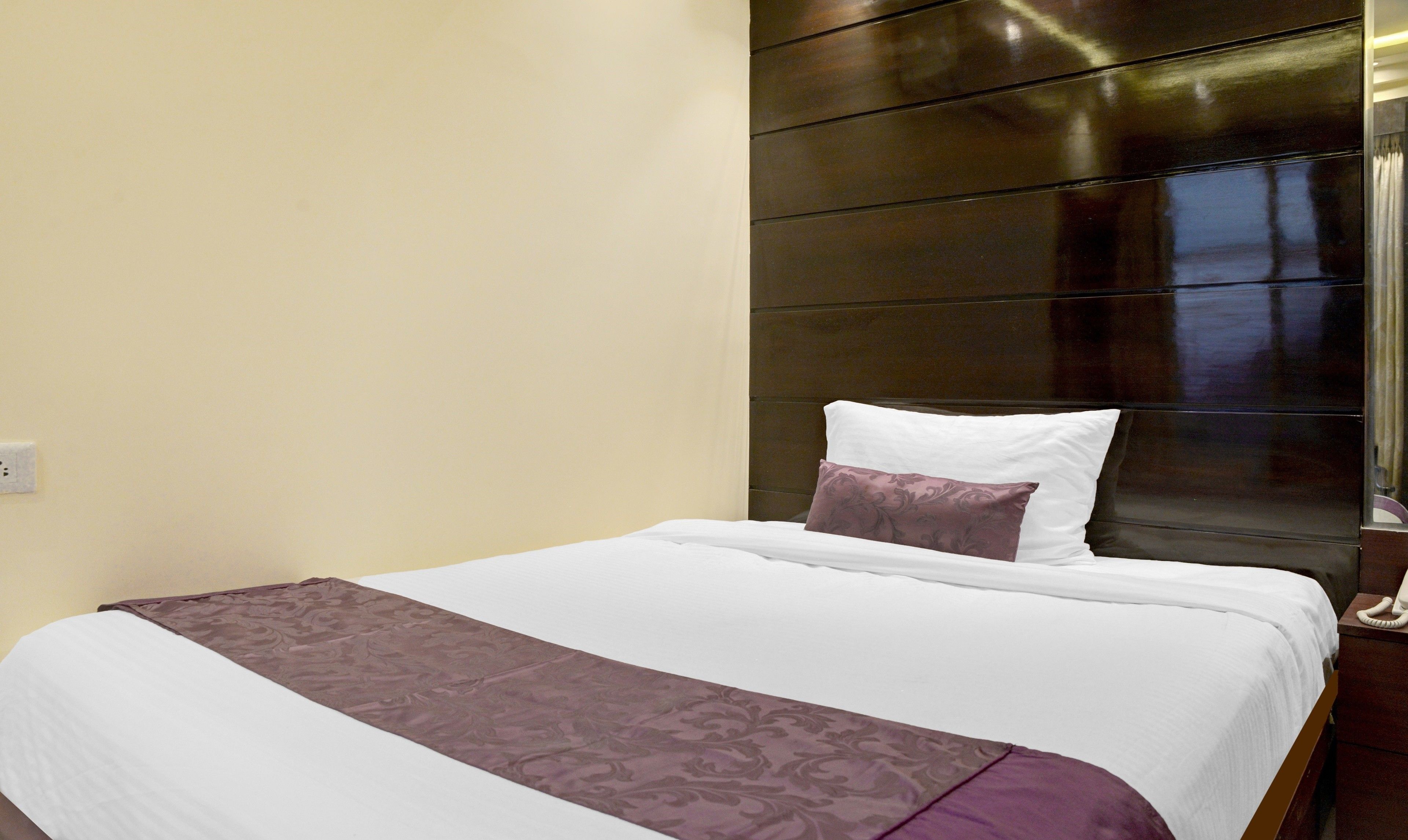 A neatly made bed with a white and purple bedspread, a wooden headboard, and a bedside telephone.