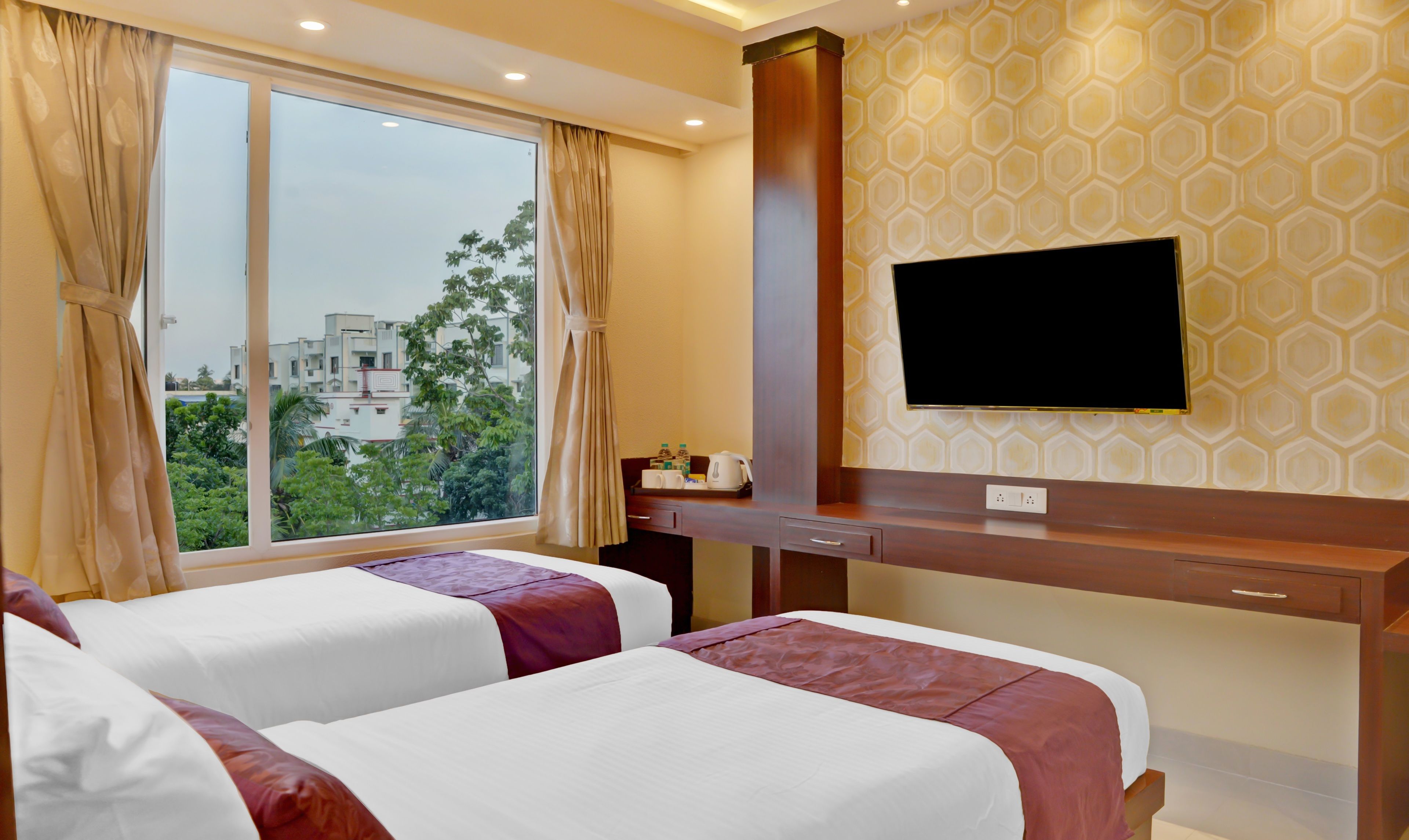 A hotel room with two beds, a desk with a flat-screen TV, and a window with curtains.