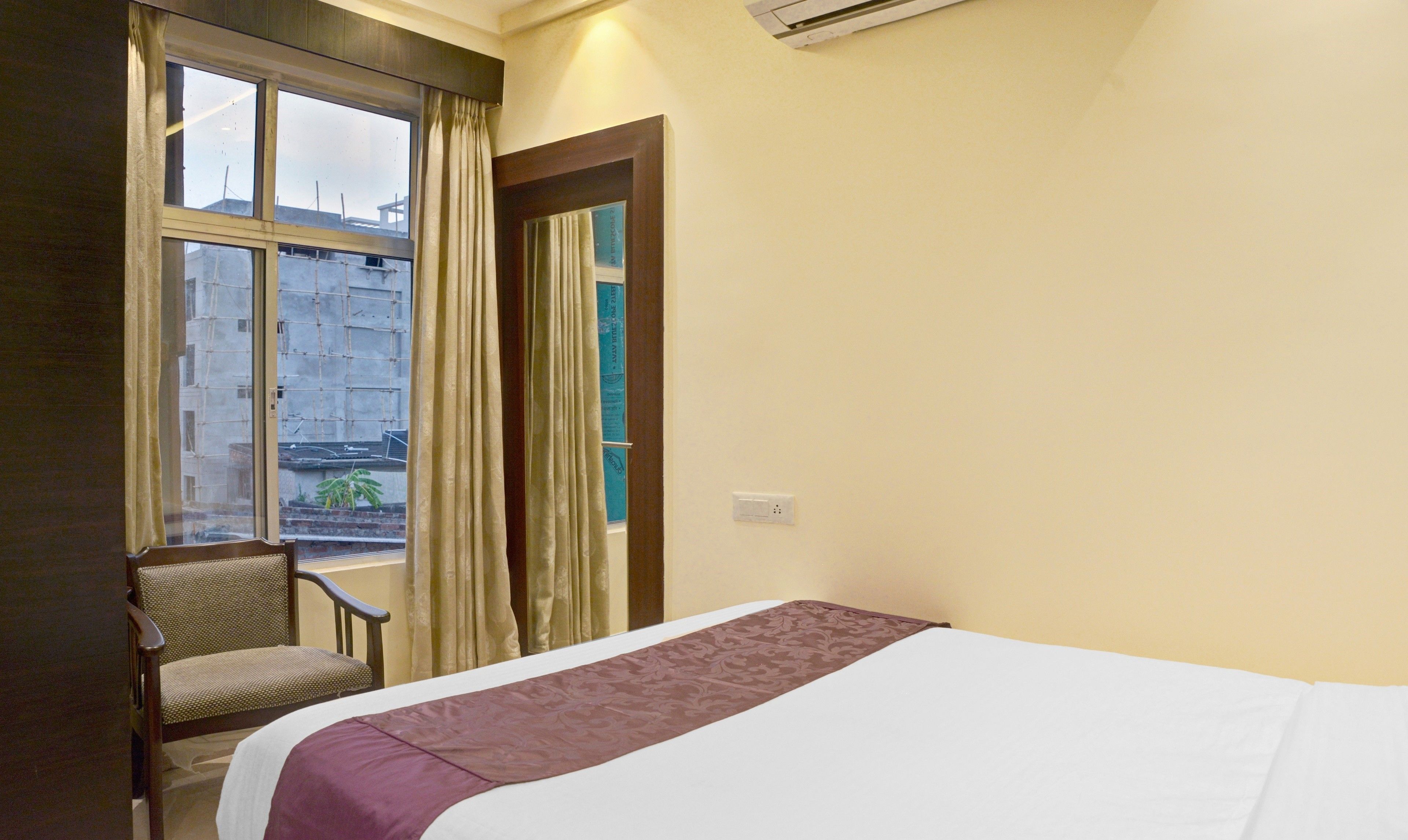 Ein Hotelzimmer mit Bett, einem Sessel, einem Fenster mit Vorhängen und Blick auf Gebäude.