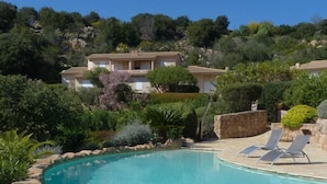 A clear blue pool with a stone border, surrounded by lush greenery and a hillside with buildings in the background. - Mini-villa No. 10 - 2/4 people (Porto-Vecchio)