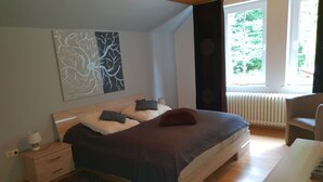Un dormitorio con cama, mesita de noche, lámpara, silla y ventana con vistas a árboles.