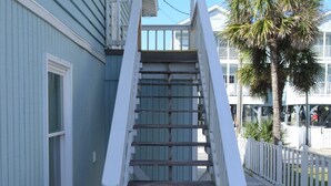 Un escalier en bois, avec des rampes blanches, mène à un porche d’un bâtiment d’un bleu clair.