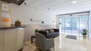 A modern reception area with a reception desk, a grey armchair, a dark sofa with cushions, and a large glass door leading outside.