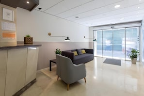 A modern reception area with a reception desk, a grey armchair, a dark sofa with cushions, and a large glass door leading outside. - Hostal Opera Ramblas (Barcelona)