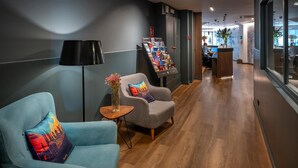 A modern lobby with a reception desk, a seating area with two armchairs, a small table with a vase, and a brochure stand.