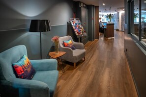 A modern lobby with a reception desk, a seating area with two armchairs, a small table with a vase, and a brochure stand.