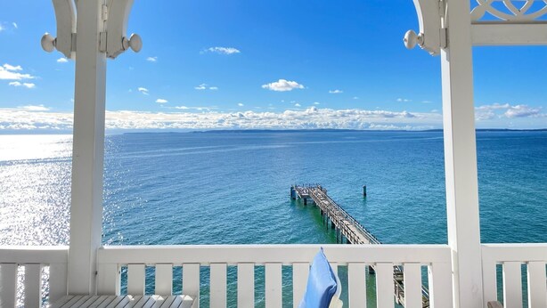 A view from a white gazebo with a wooden deck, overlooking a calm sea and a pier extending into the water.