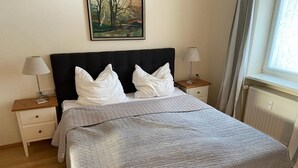 A neatly made bed with a quilted blanket, two bedside tables with lamps, a painting on the wall, and a window with curtains.