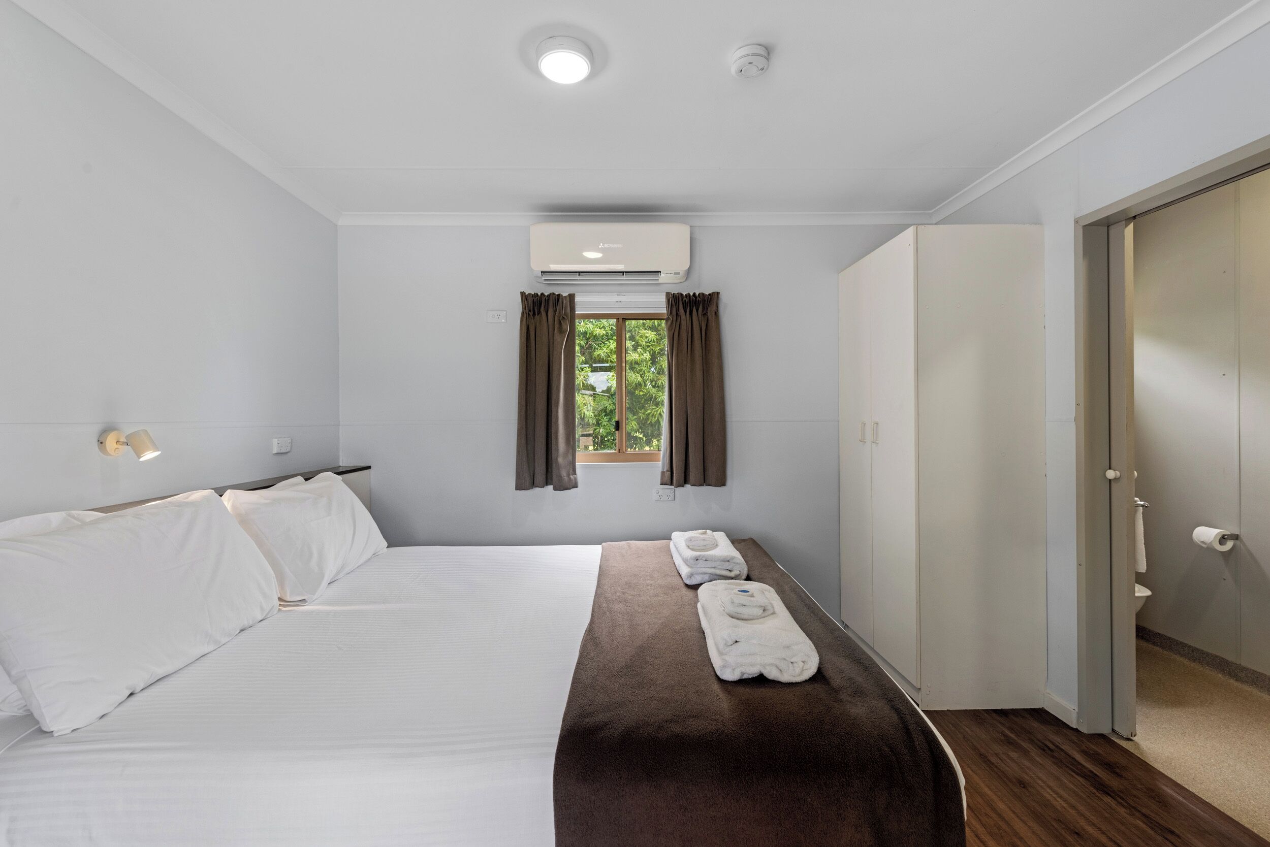 A hotel room with a bed, a window with curtains, an air conditioning unit, a towel rack, and a door to the bathroom.