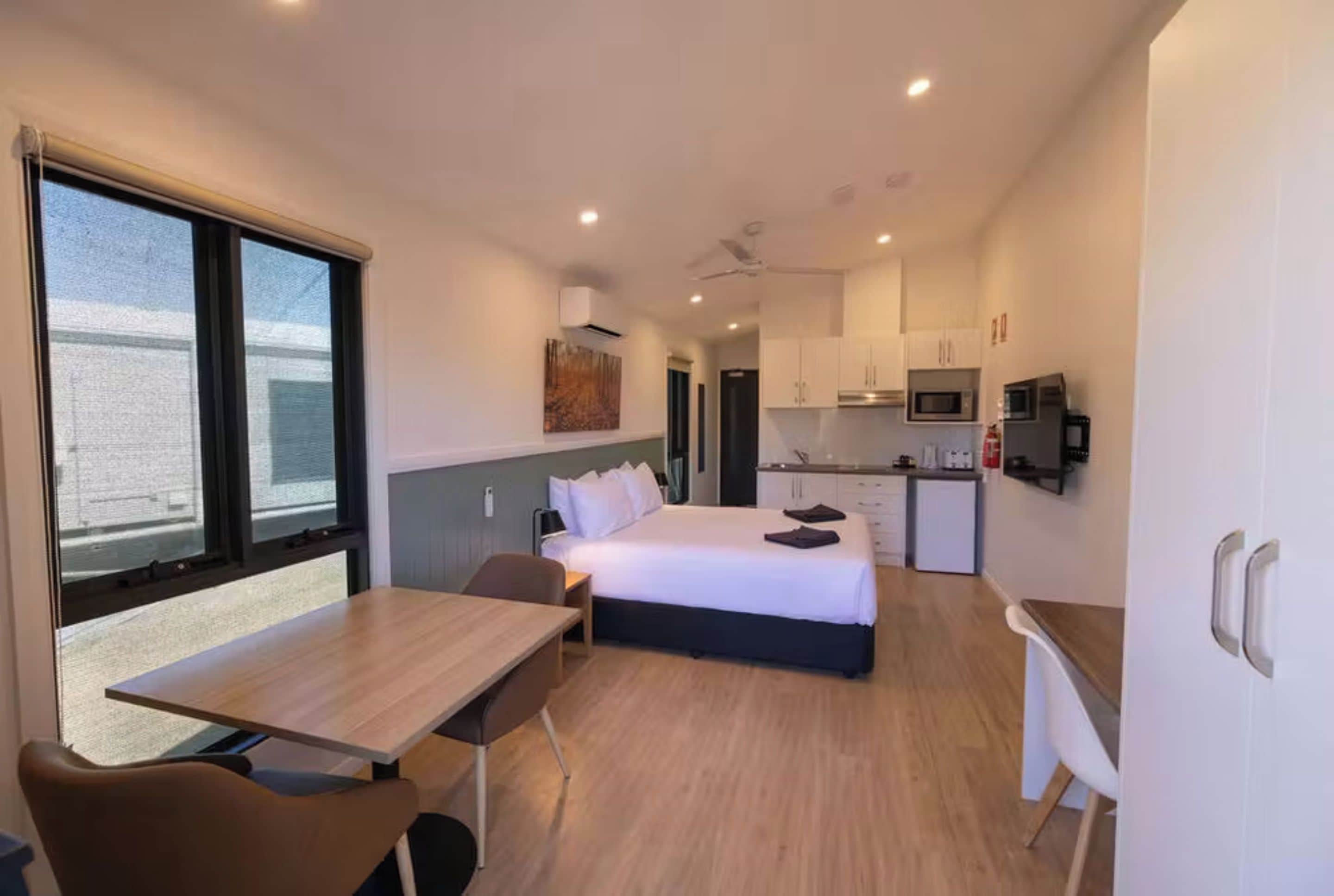 A compact hotel room with a bed, a desk, a chair, a kitchenette, and a large window.