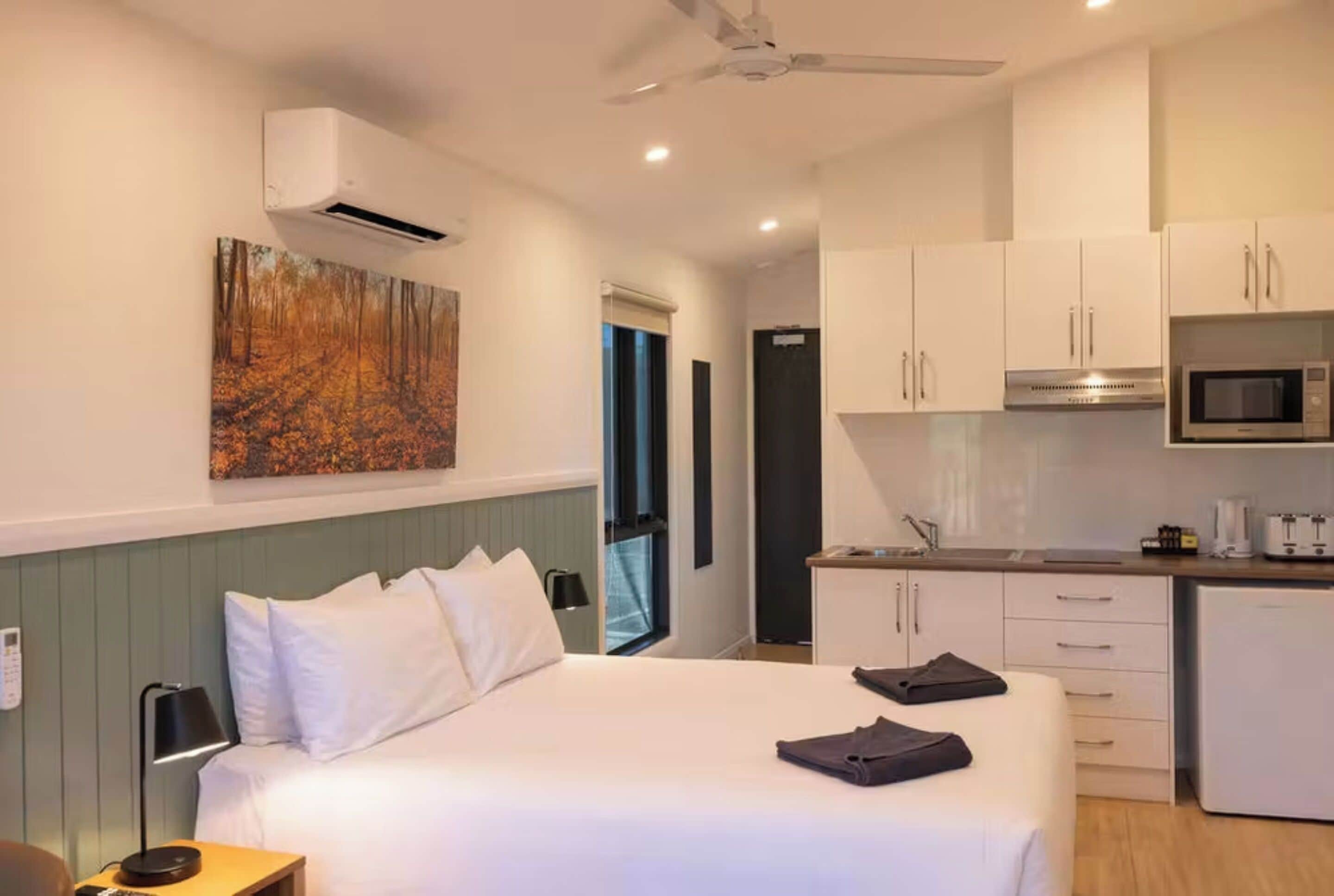 Quarto de hotel com cama, escrivaninha, cozinha compacta e um quadro na parede.