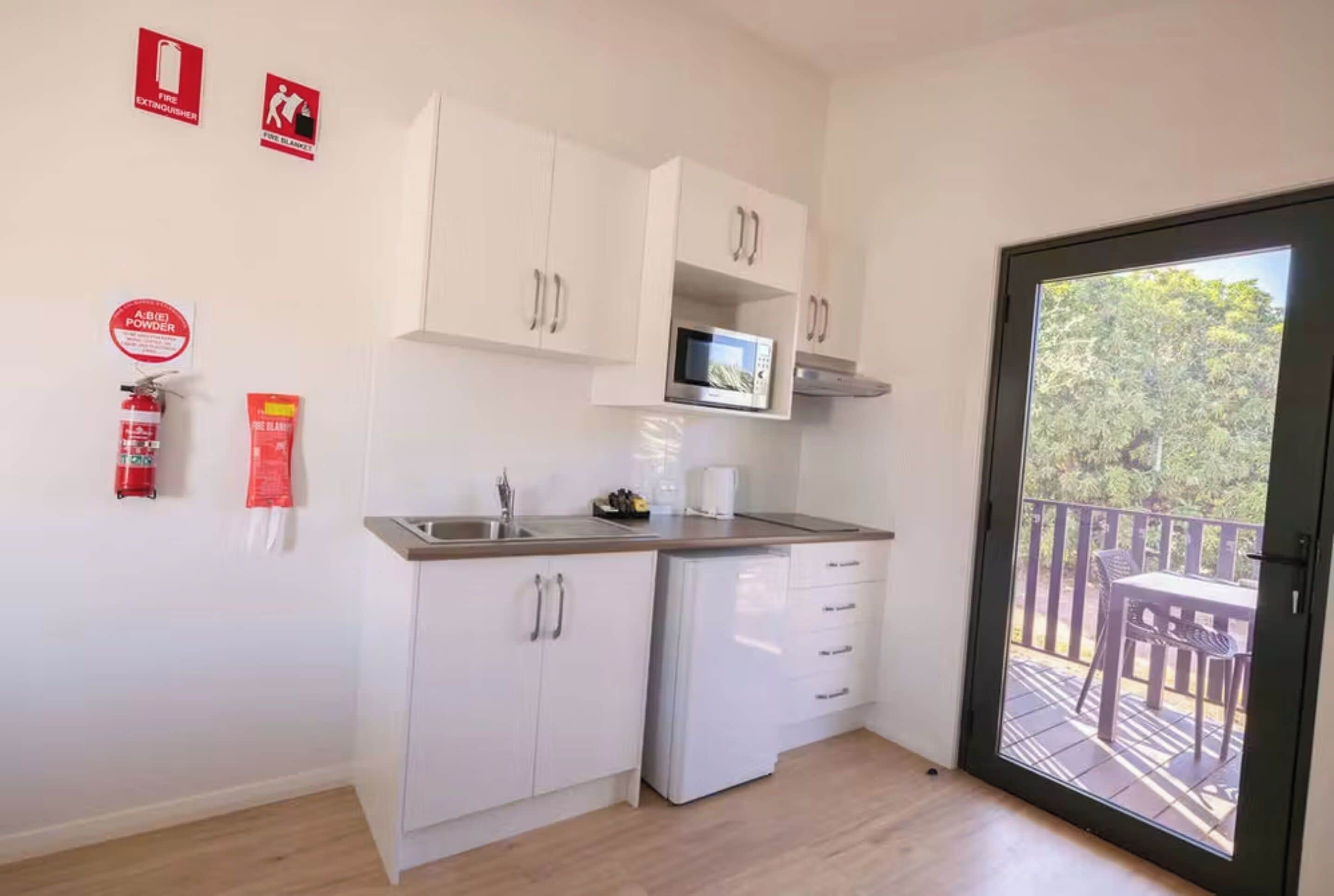 A compact kitchen with white cabinets, a microwave, and a sink. There are fire safety signs on the wall, and a balcony with outdoor furniture is visible through the door.
