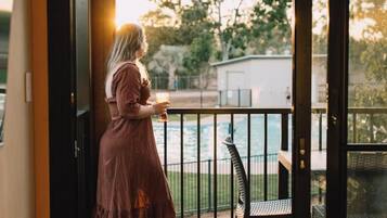 A woman in a brown dress standing by a pool, holding a drink, with a view of the pool and a building in the background.