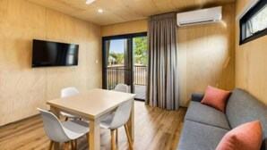 Una habitación moderna con suelo de madera, un televisor de pantalla plana montado en la pared, una mesa de comedor con sillas, un sofá gris con cojines y un ventanal corredero que da a un espacio exterior.