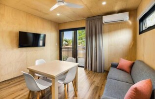 A modern room with a wooden floor, a flat-screen TV mounted on the wall, a dining table with chairs, a grey sofa with cushions, and a sliding glass door leading to an outdoor area.