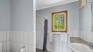 A bathroom with white tiles, a shower, a sink, and a window with a view of greenery.