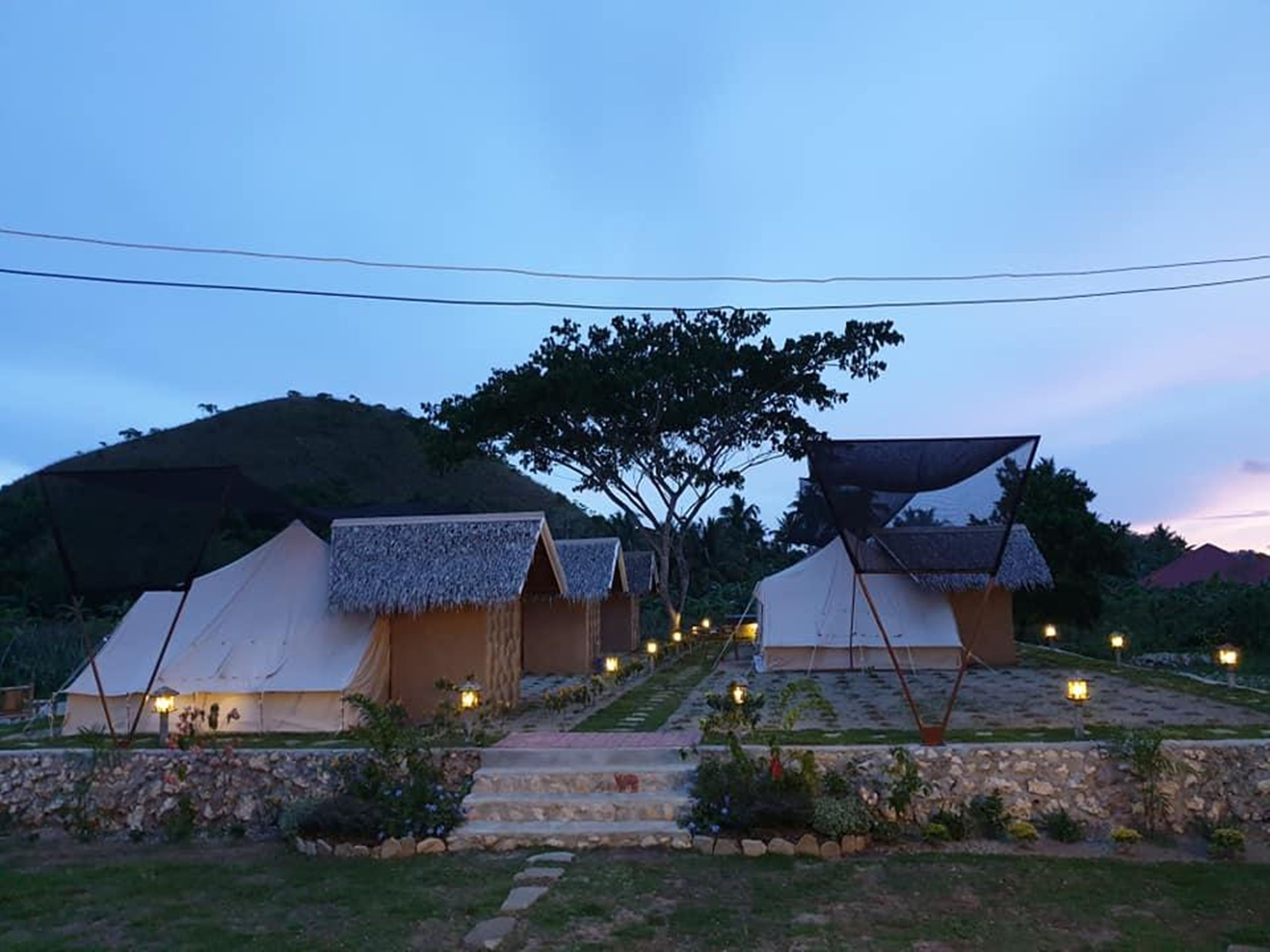 A row of thatched-roof tents with a stone pathway leading to them, set against a hilly backdrop.
