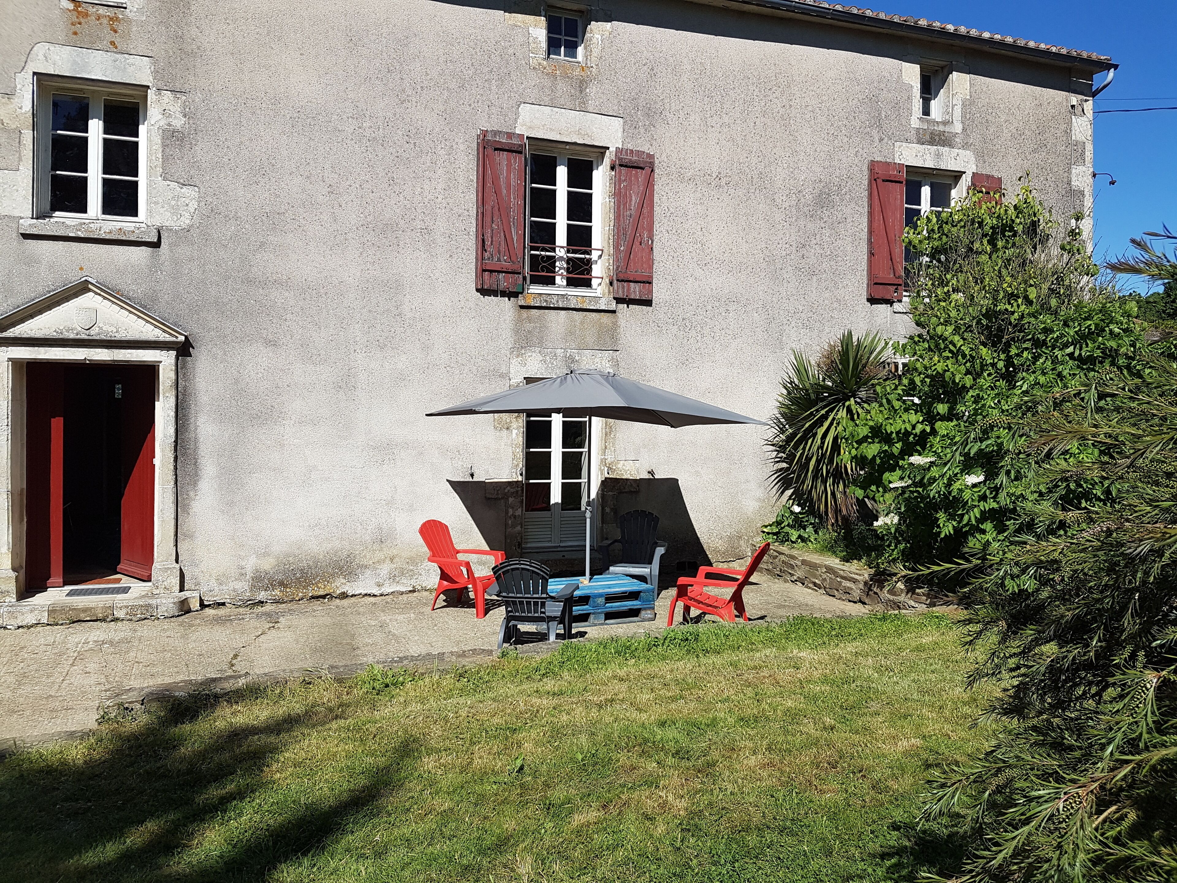 Un bâtiment à deux étages, doté d’une façade en pierre, de volets rouges et d’un petit porche avec des sièges extérieurs.