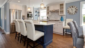 A modern kitchen with a central island, white bar stools, and a large clock on the wall. - Luxury Monument Beach Villa /Short term rentals (Bourne)