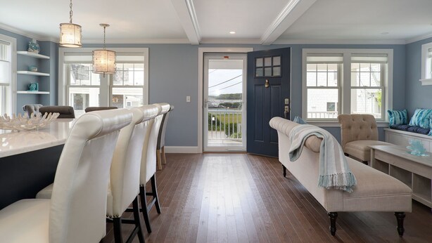 A spacious room with a wooden floor, a bar area with white bar stools, a cushioned bench, and a door leading outside. - Luxury Monument Beach Villa /Short term rentals (Bourne)
