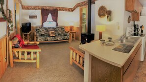 Ein gemütliches Blockhaus-Interieur mit einer Couch, einem kleinen Esstisch, einer Küchenzeile mit Herd und Spüle sowie einem Kamin.