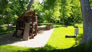 Ein Spielplatz mit Rutsche und Klettergerüsten, umgeben von Bäumen und einem Schild, das zum See weist.