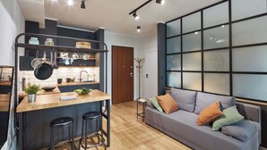 A modern kitchen with a bar area, a grey sofa, and a large window with blinds. - Modern Apartment with Balcony | Close to the Metro (Warszawa)