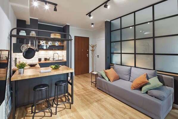 A modern kitchen with a bar area, a grey sofa, and a large window with blinds.