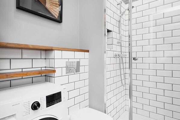 Un baño moderno con azulejos blancos tipo metro, un estante de madera, una lavadora y una ducha con mampara de vidrio.
