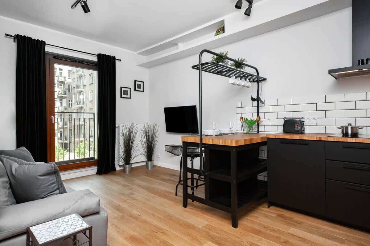 Una cocina moderna con encimera de madera, armarios negros y una estantería colgante negra. Hay una mesa de comedor con una silla, un sofá y un televisor montado en la pared.