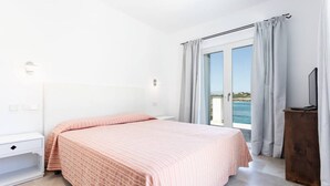 Un dormitorio con cama, mesita de noche, cómoda de madera y vista al mar a través de una puerta corrediza de vidrio.