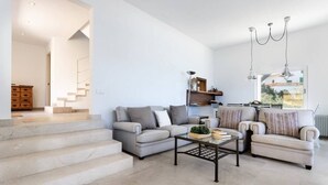 Una sala de estar moderna con un sofá, sillones y una mesa de centro. Se ve una escalera que lleva a otra habitación, y una ventana con vistas al exterior.