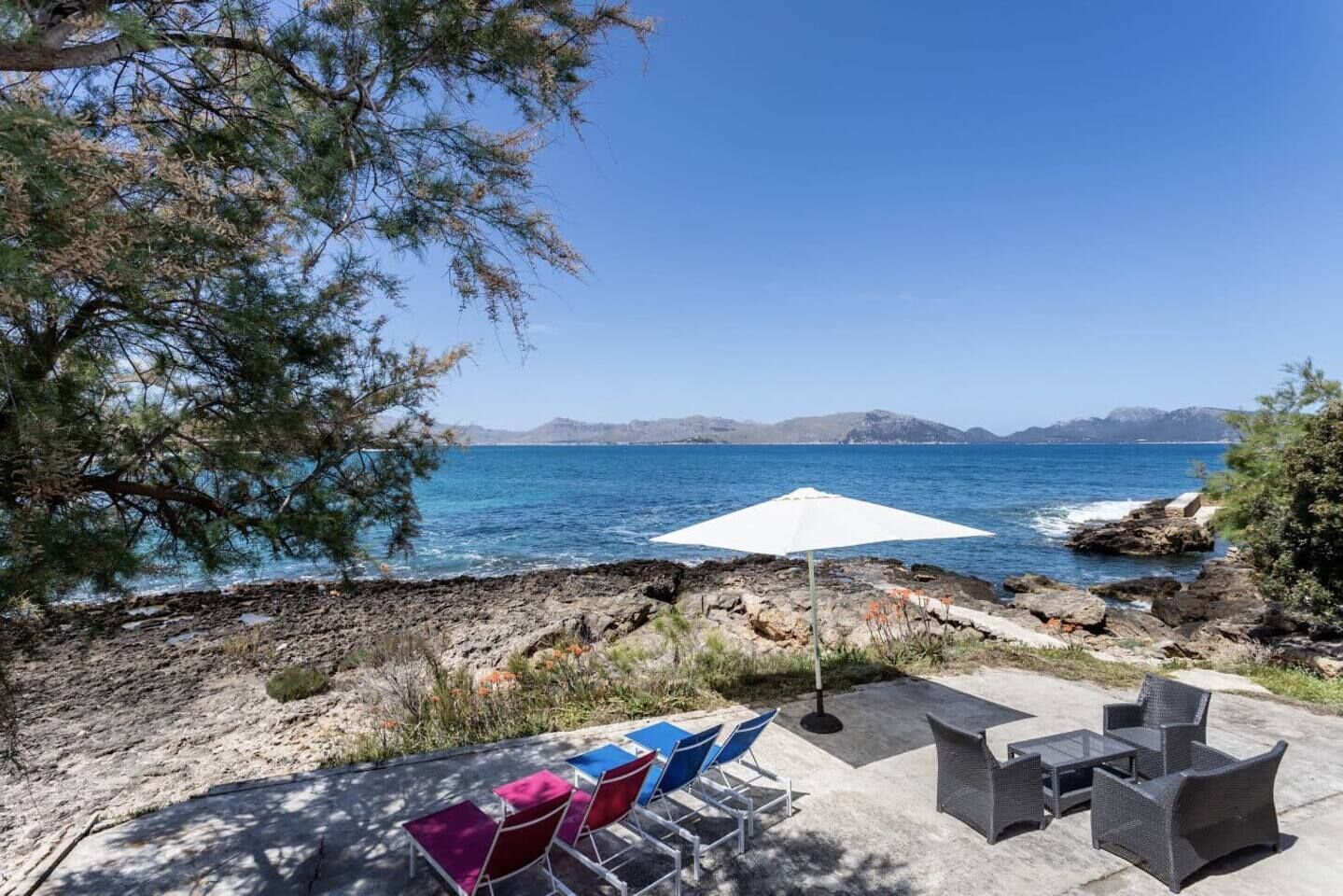 A patio with outdoor furniture, a white umbrella, and a view of the sea and mountains.