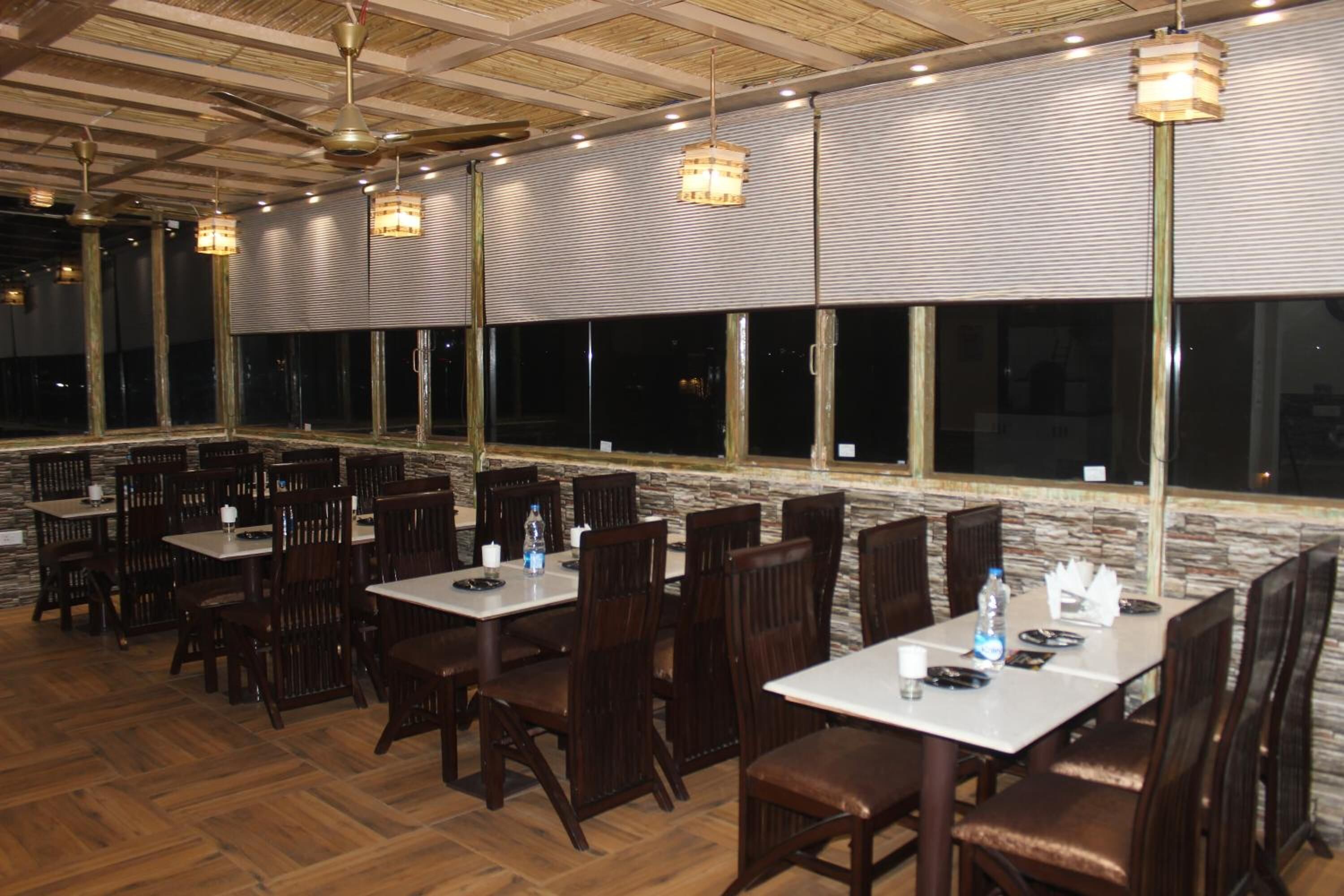Un restaurante con varias mesas y sillas, ventanas amplias y lámparas colgantes.