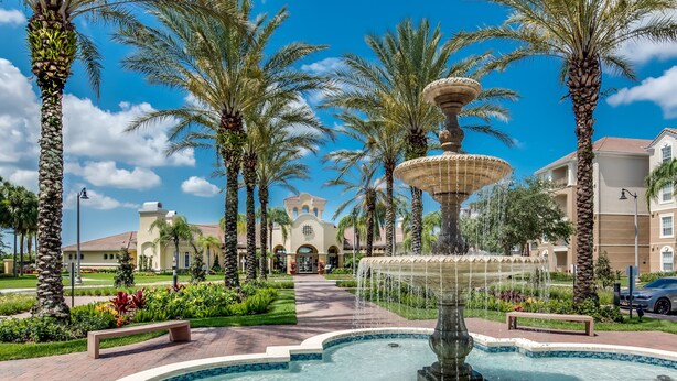 A fountain surrounded by palm trees and a pathway leading to a building with arched entrances.
