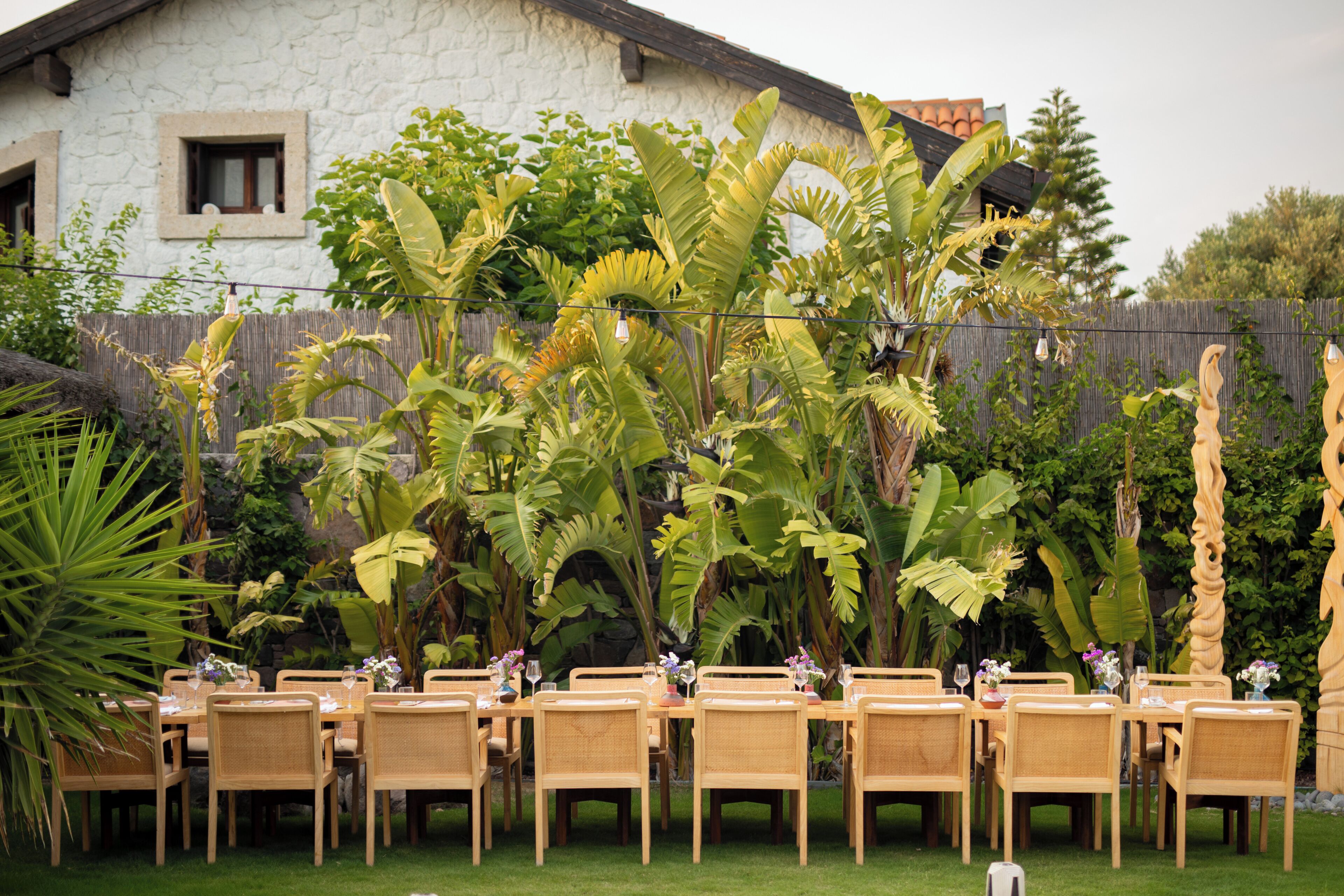 Un'area pranzo all'aperto con tavoli e sedie in legno, circondata da rigogliosa vegetazione tropicale e un edificio in pietra sullo sfondo.