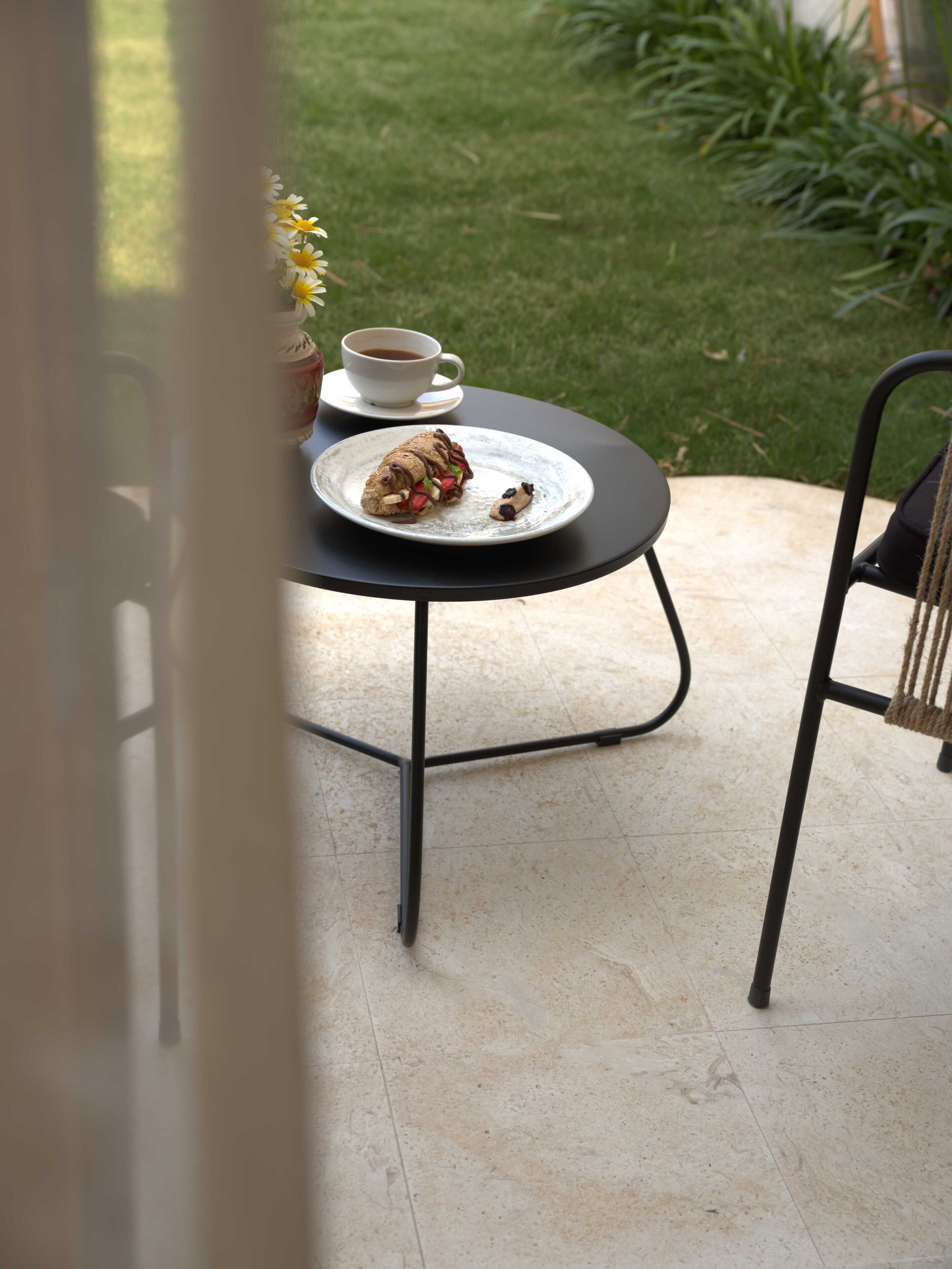 A round table with a plate of food and a cup of coffee, a chair, and a vase with flowers.