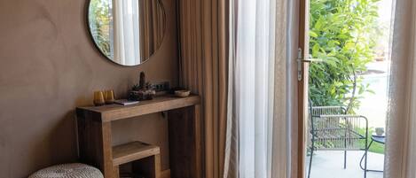 A room with a wooden console table, a round mirror, a small stool, and a view of a garden through a glass door.