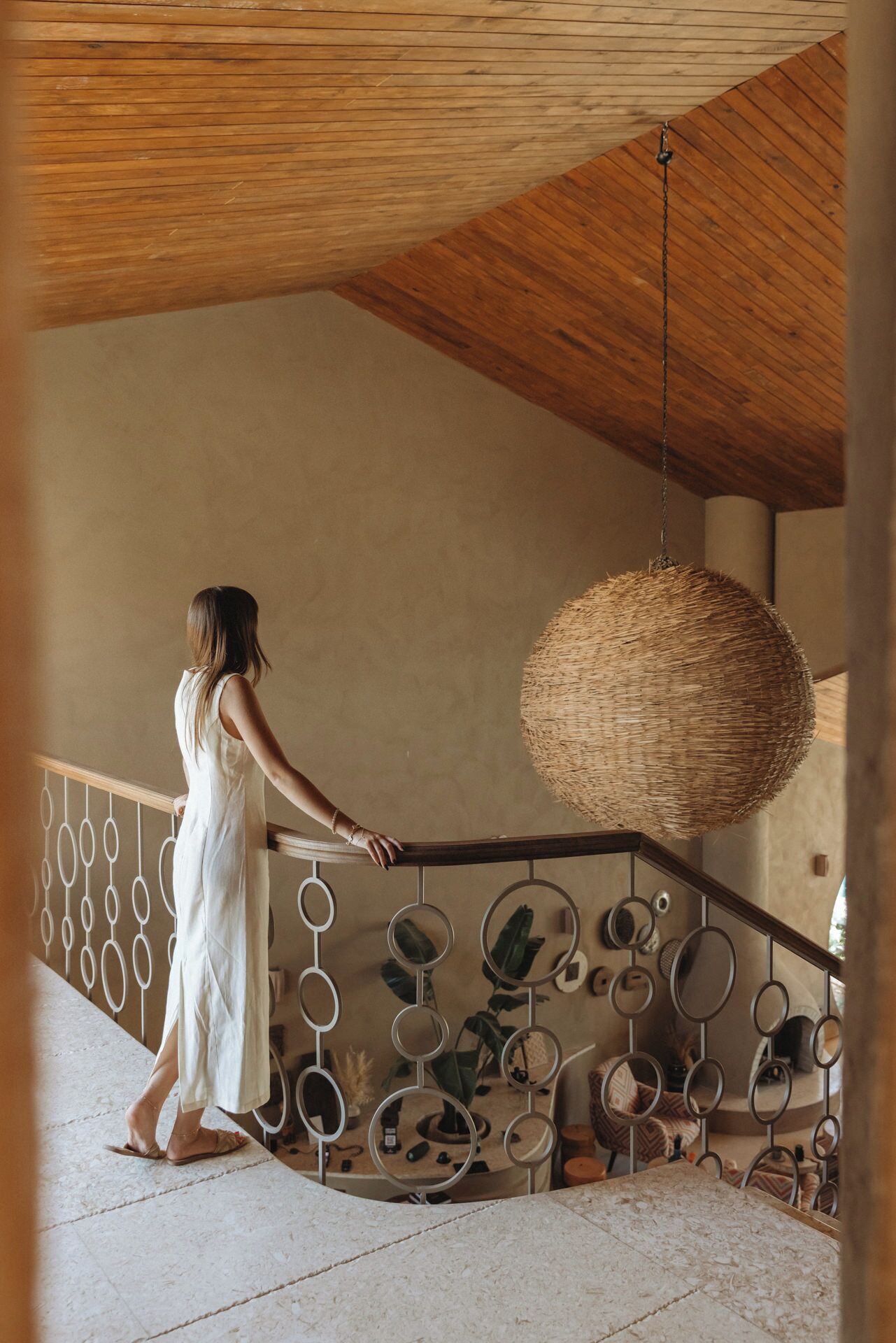 A woman in a white dress standing on a balcony with a wooden ceiling and a woven light fixture.