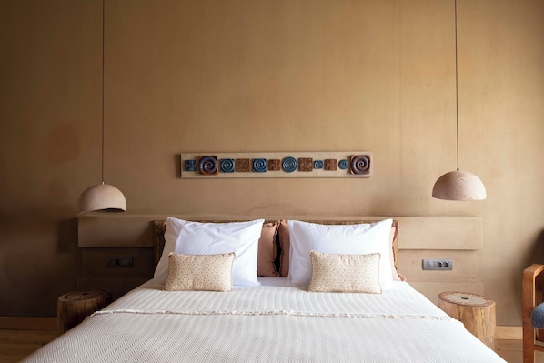Una cama grande con una colcha blanca texturizada, dos almohadas y un cabecero con un panel decorativo en la parte superior. Hay dos mesitas de noche de madera con lámparas a ambos lados de la cama.