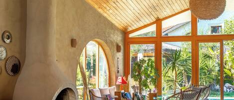 A spacious living area with a high wooden ceiling, a fireplace, comfortable seating, and large windows offering a view of the outdoors.