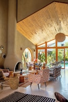 Un amplio salón con techo de madera alto, chimenea, asientos cómodos y grandes ventanales con vistas al exterior.
