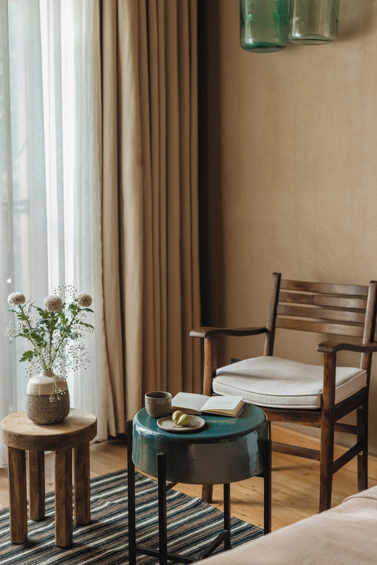 A cozy room with a wooden chair, a small round table with a book and a cup, and a vase with flowers on a wooden stool.