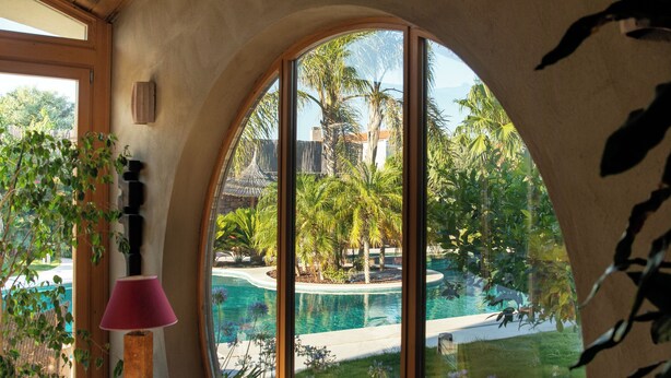 A room with a large curved window, wooden ceiling, and a view of a swimming pool and tropical landscape. - Pachamama Alacati  (Cesme)