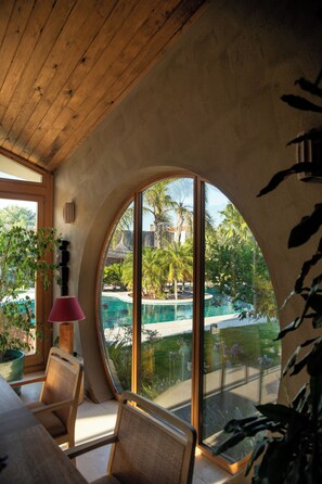 Una habitación con un amplio ventanal curvo, techo de madera y vistas a una piscina y paisaje tropical.