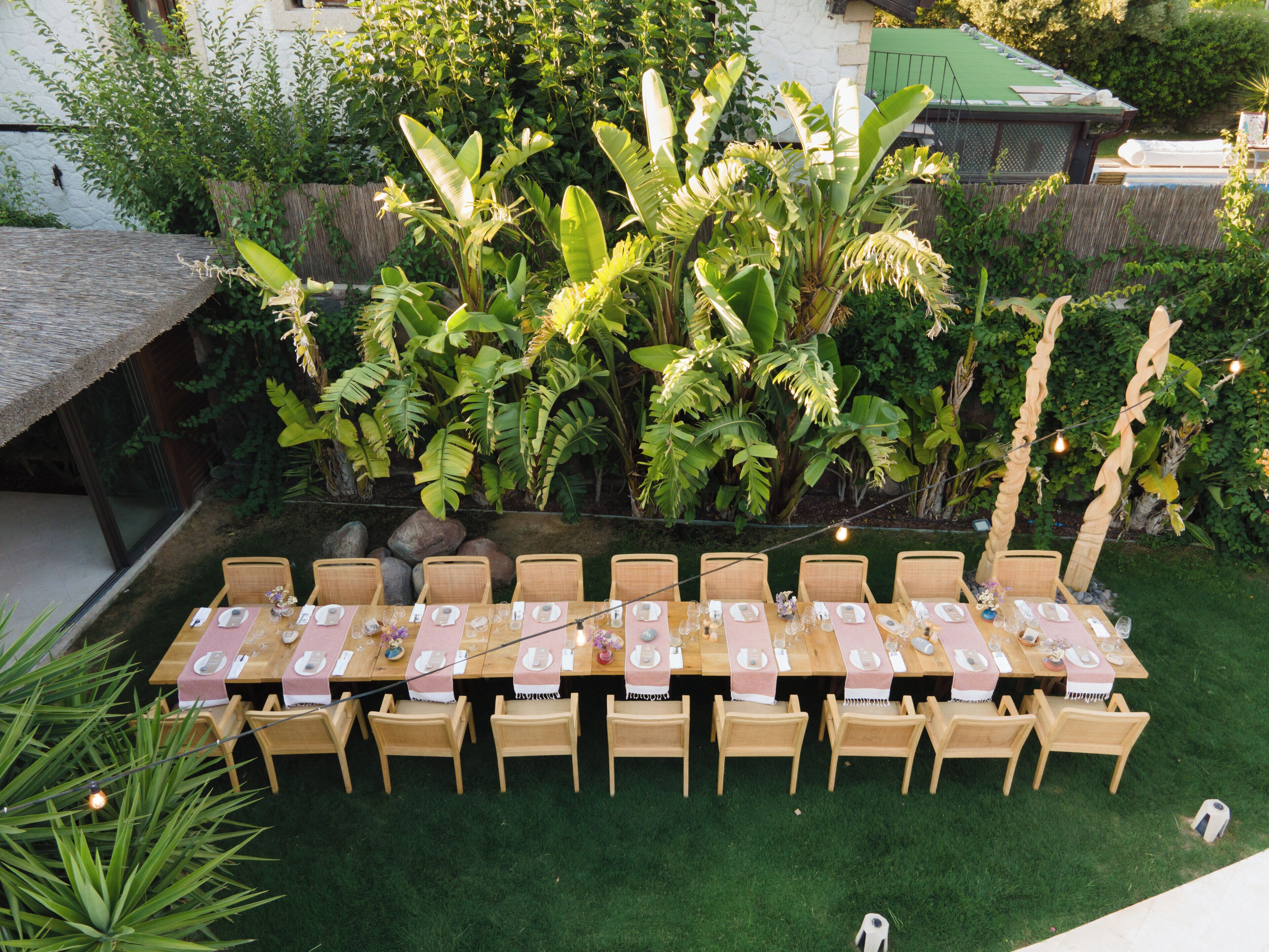 An outdoor dining area with a long table set for a meal, surrounded by chairs, and a backdrop of lush greenery and a building.