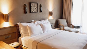 Una habitación de hotel con una cama grande, cabecero de madera, mesitas de noche con lámparas, una silla y una pequeña mesa con una cesta y papeles.